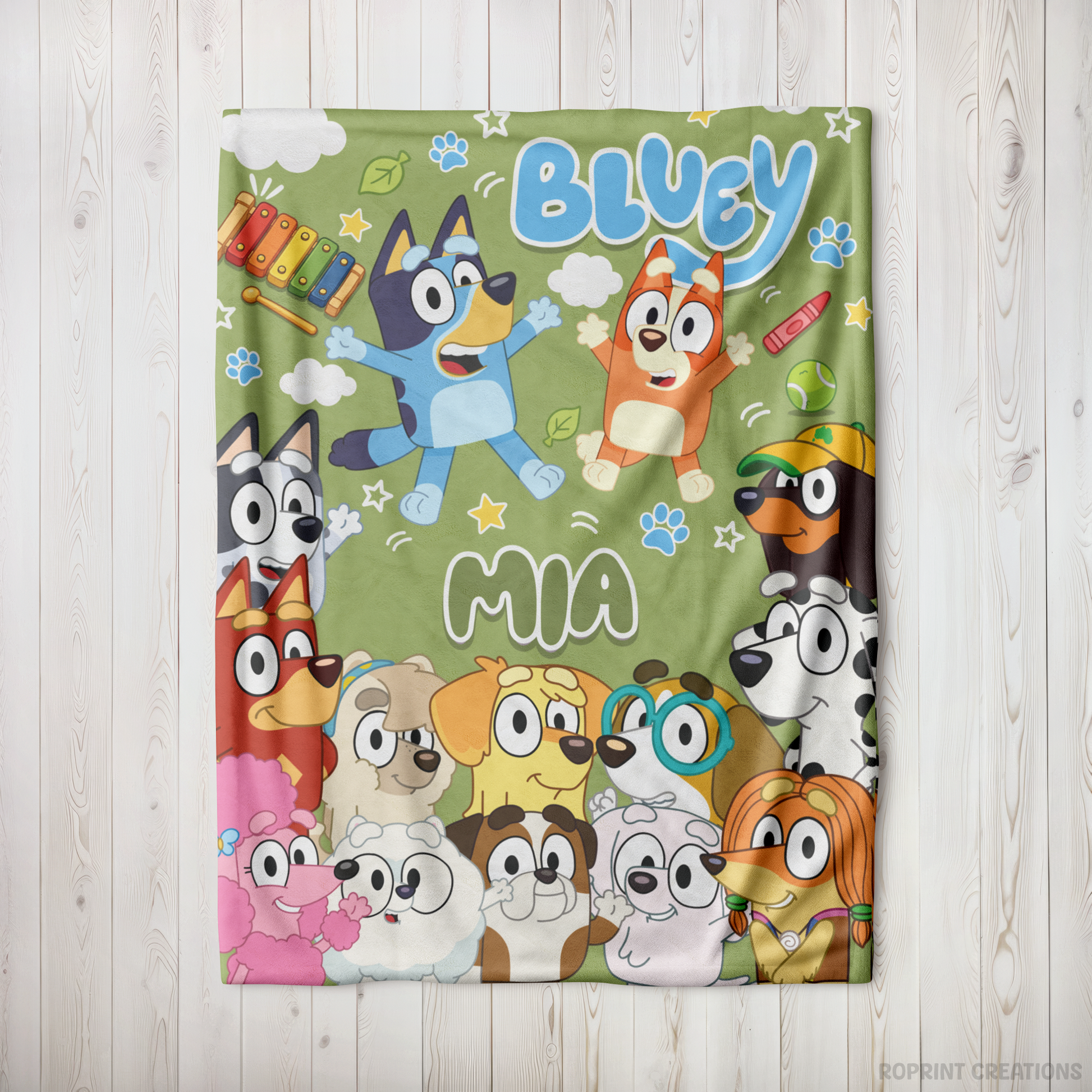 A personalized Bluey-themed fleece blanket featuring characters Bluey, Bingo, and other pups, with the names 'BLUEY' and 'MIA' printed on a light green background, displayed flat on a white wood panel surface.