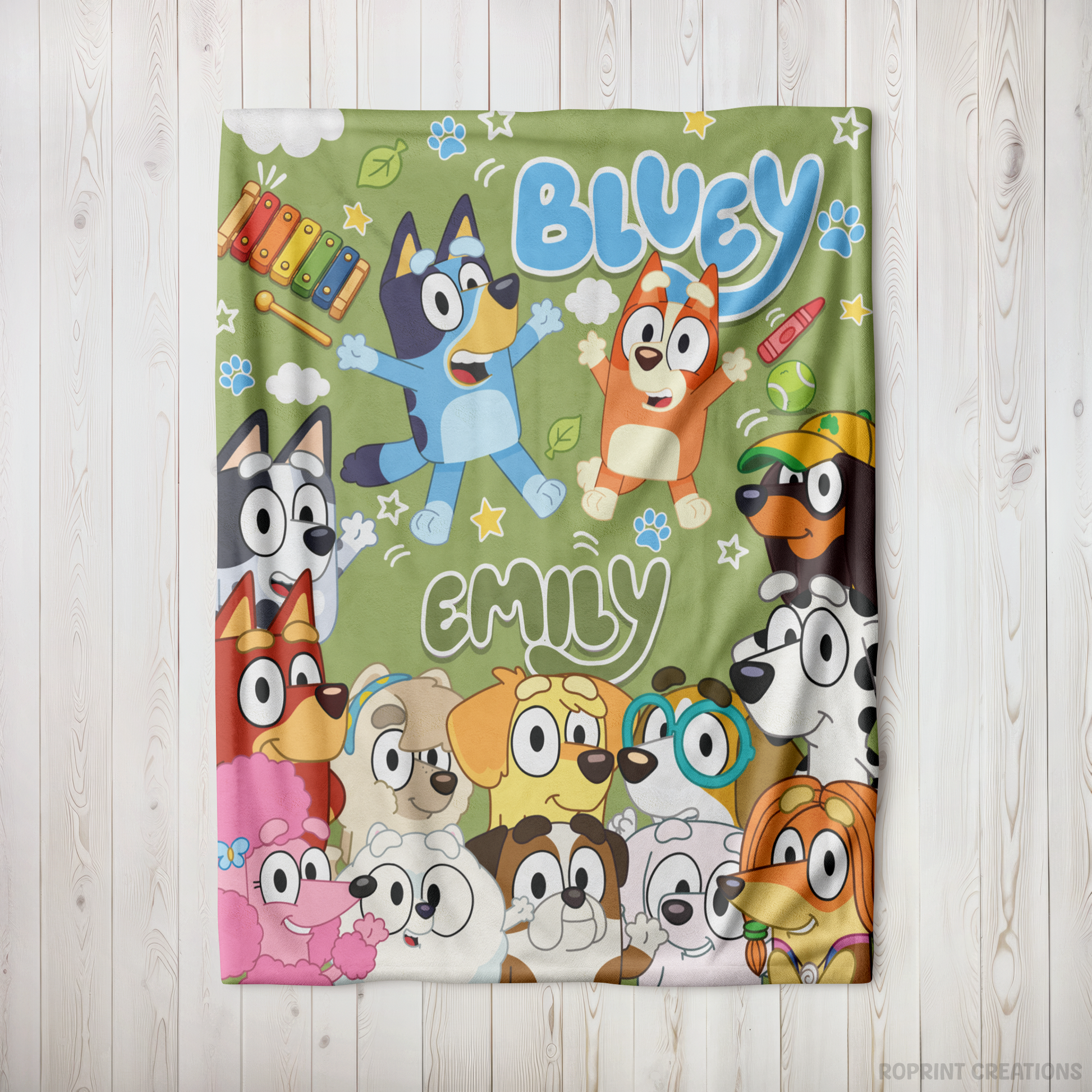 A personalized Bluey-themed blanket featuring cartoon dog characters and the names 'Bluey' and 'Emily' printed on a light olive green background, displayed flat on a white wood plank surface.