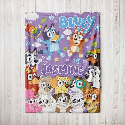 A personalized Bluey-themed fleece blanket featuring cartoon characters and the names 'BLUEY' and 'JASMINE' printed on a purple background, displayed flat on a light wood plank surface.