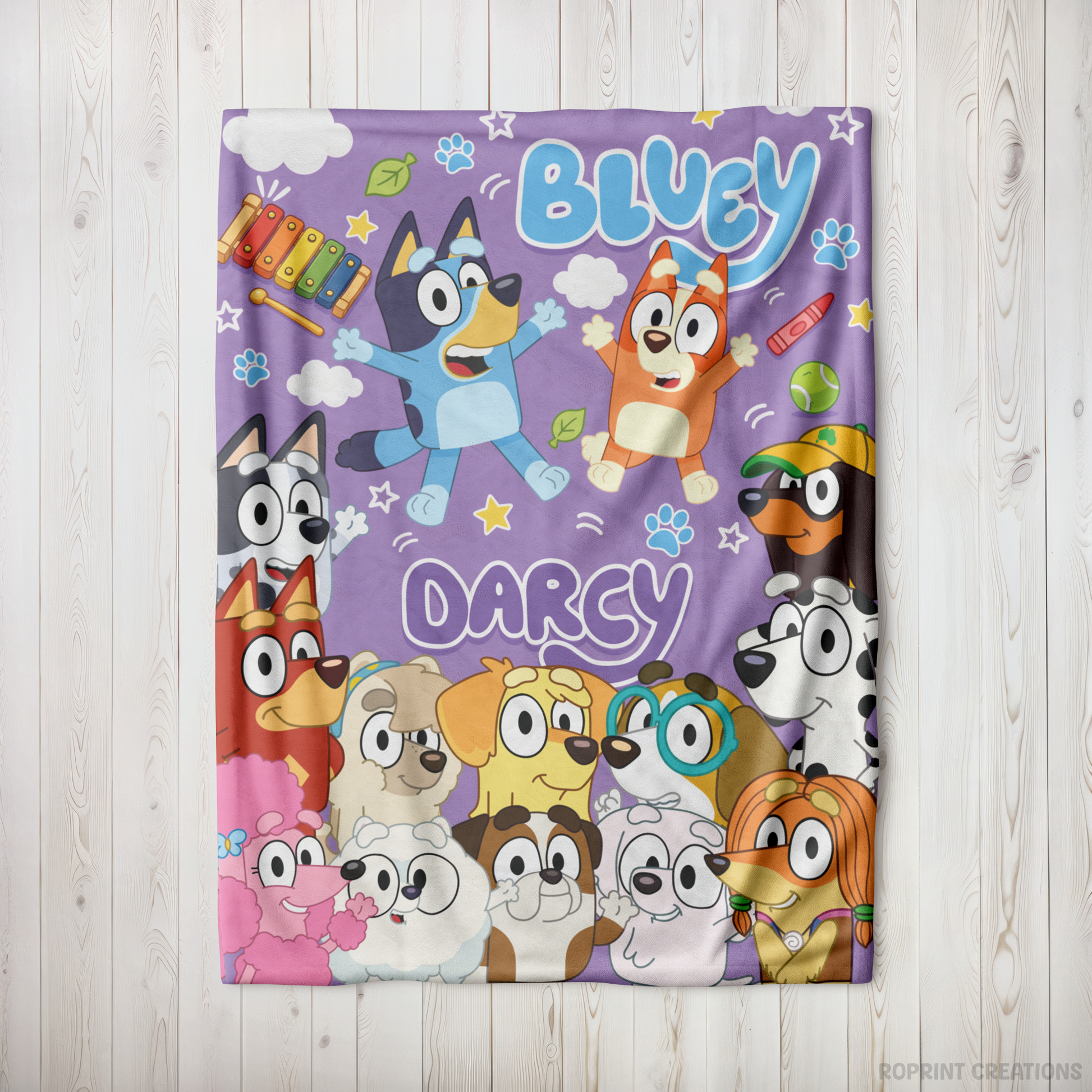 A personalized Bluey-themed blanket featuring cartoon characters Bluey, Darcy, and other pups on a purple background, laid flat on a light wood plank surface.