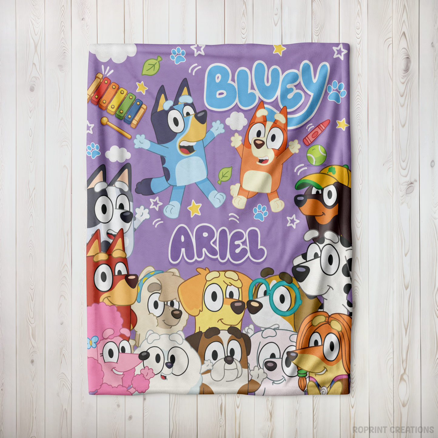 A personalized Bluey-themed blanket featuring characters Bluey, Angel, and other dog friends on a purple background, displayed flat on a light wood plank surface.