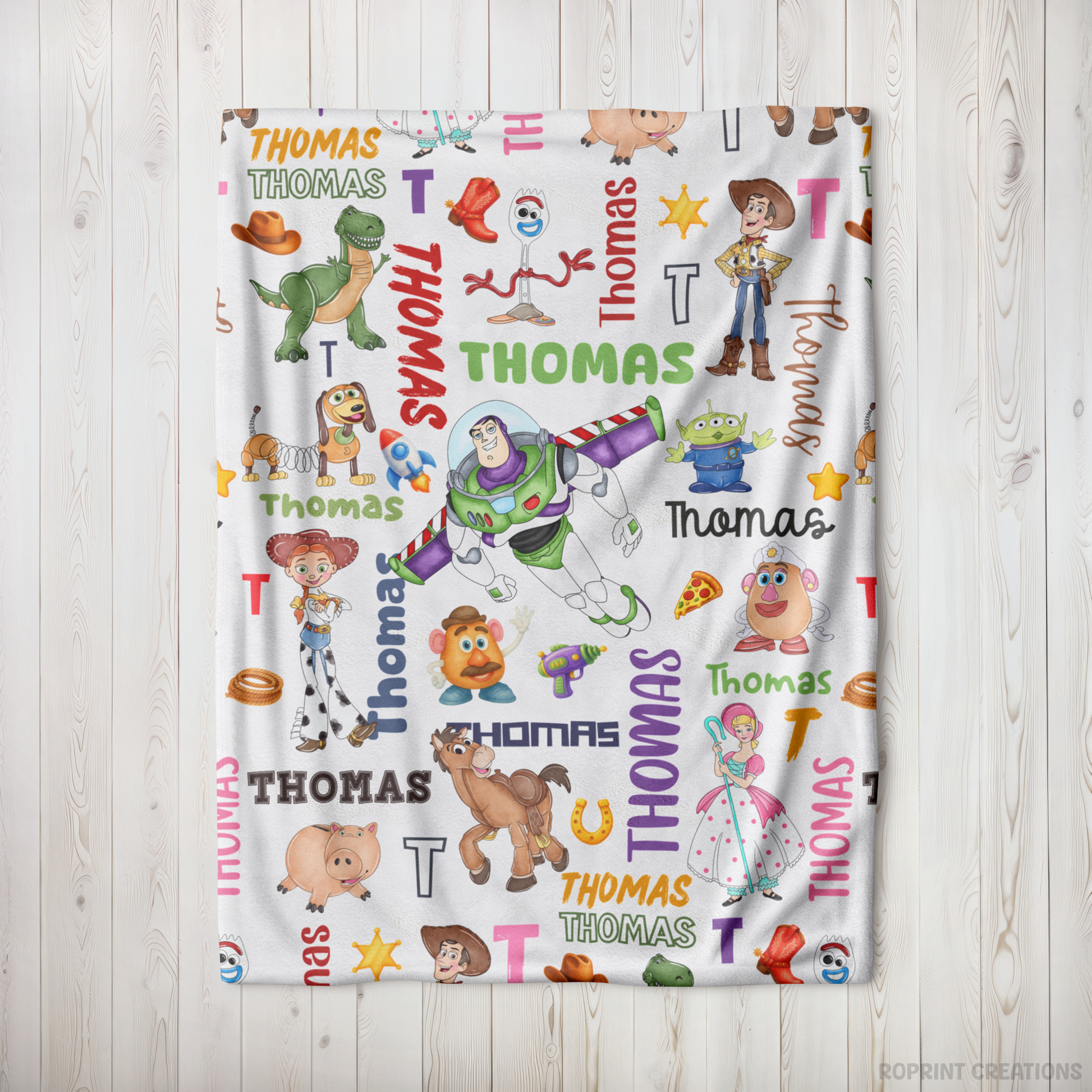 A white personalized blanket featuring a colorful repeating pattern of Toy Story characters, names, and letters, displayed flat against a light wood panel background.