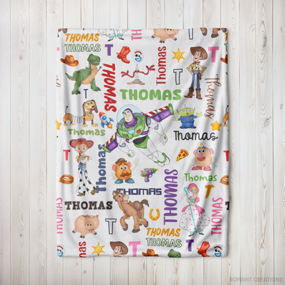 A white personalized blanket featuring a colorful repeating pattern of Toy Story characters, names, and letters, displayed flat against a light wood panel background.