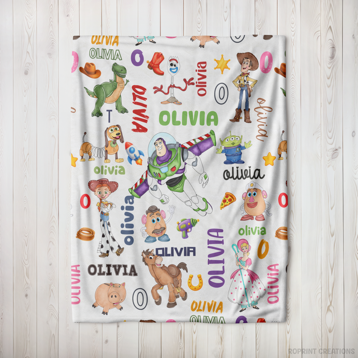 A personalized blanket featuring colorful Toy Story character illustrations and the name 'OLIVIA' repeated across a white background, displayed flat against a light wood plank wall.
