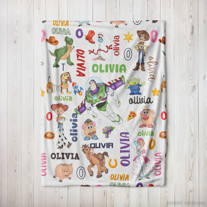 A personalized blanket featuring colorful Toy Story character illustrations and the name 'OLIVIA' repeated across a white background, displayed flat against a light wood plank wall.